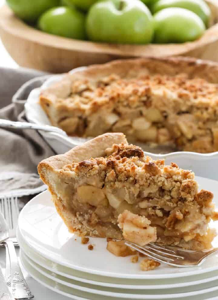 A white pie dish with a slice of apple crumble pie, also known as a dutch apple pie with a crumbled topping and green apples in the background in a wood bowl.