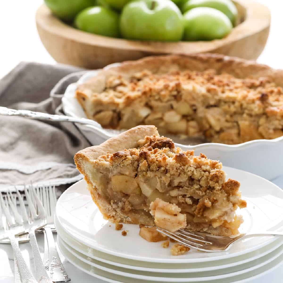 A white pie dish with a slice of apple crumble pie, also known as a dutch apple pie with a crumbled topping and green apples in the background in a wood bowl.