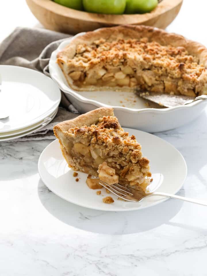 A white pie dish with a slice of apple crumble pie, also known as a dutch apple pie with a crumbled topping and green apples in the background in a wood bowl.