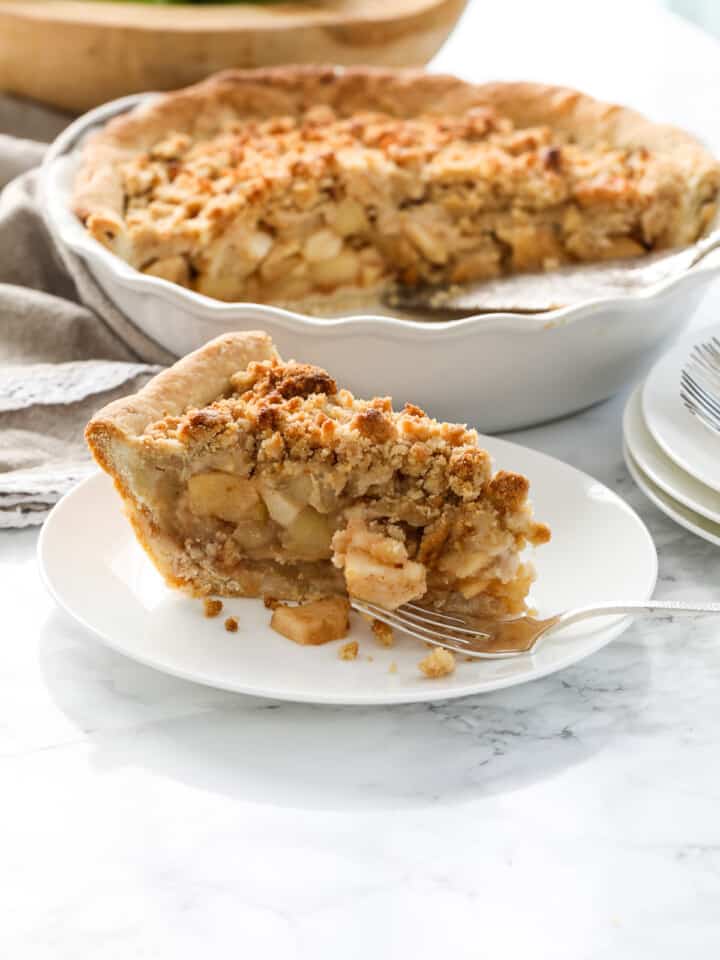 A large piece of apple crumble pie on a white plate with a silver fork and the pie plate with half a pie left in the background.