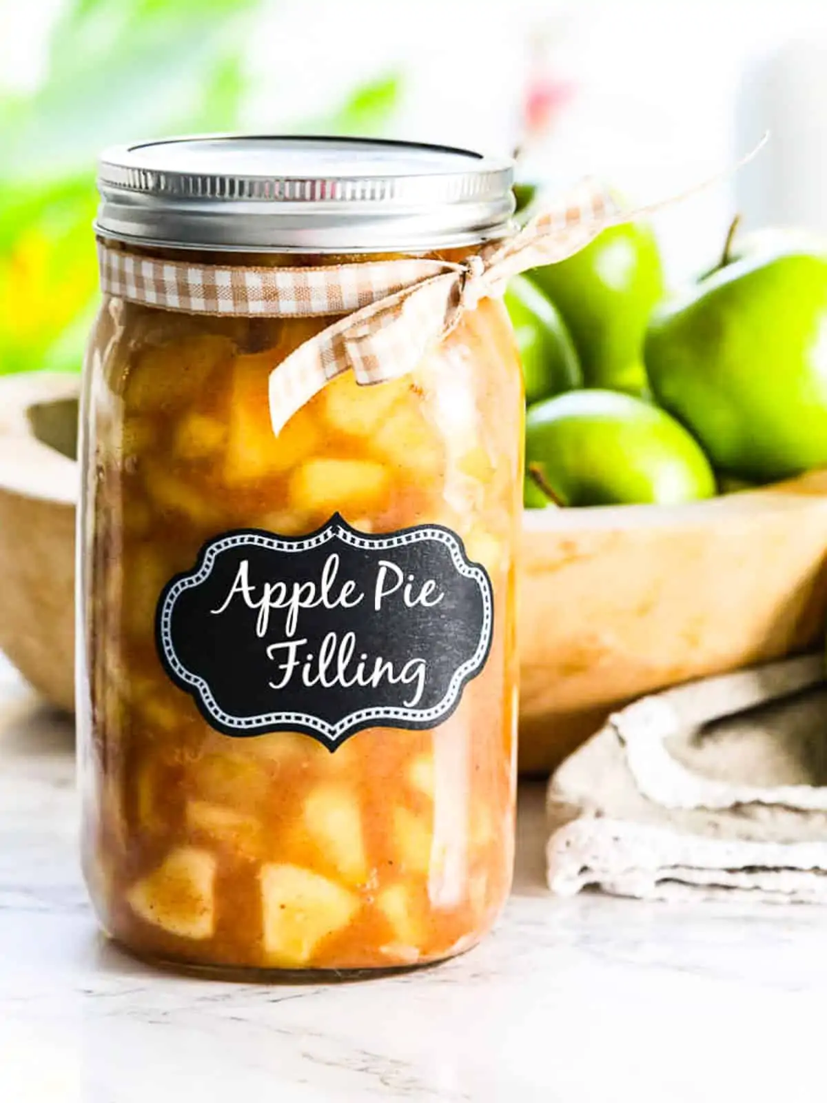 A large glass jar filled with apple pie filling and wood bowl of green apples.