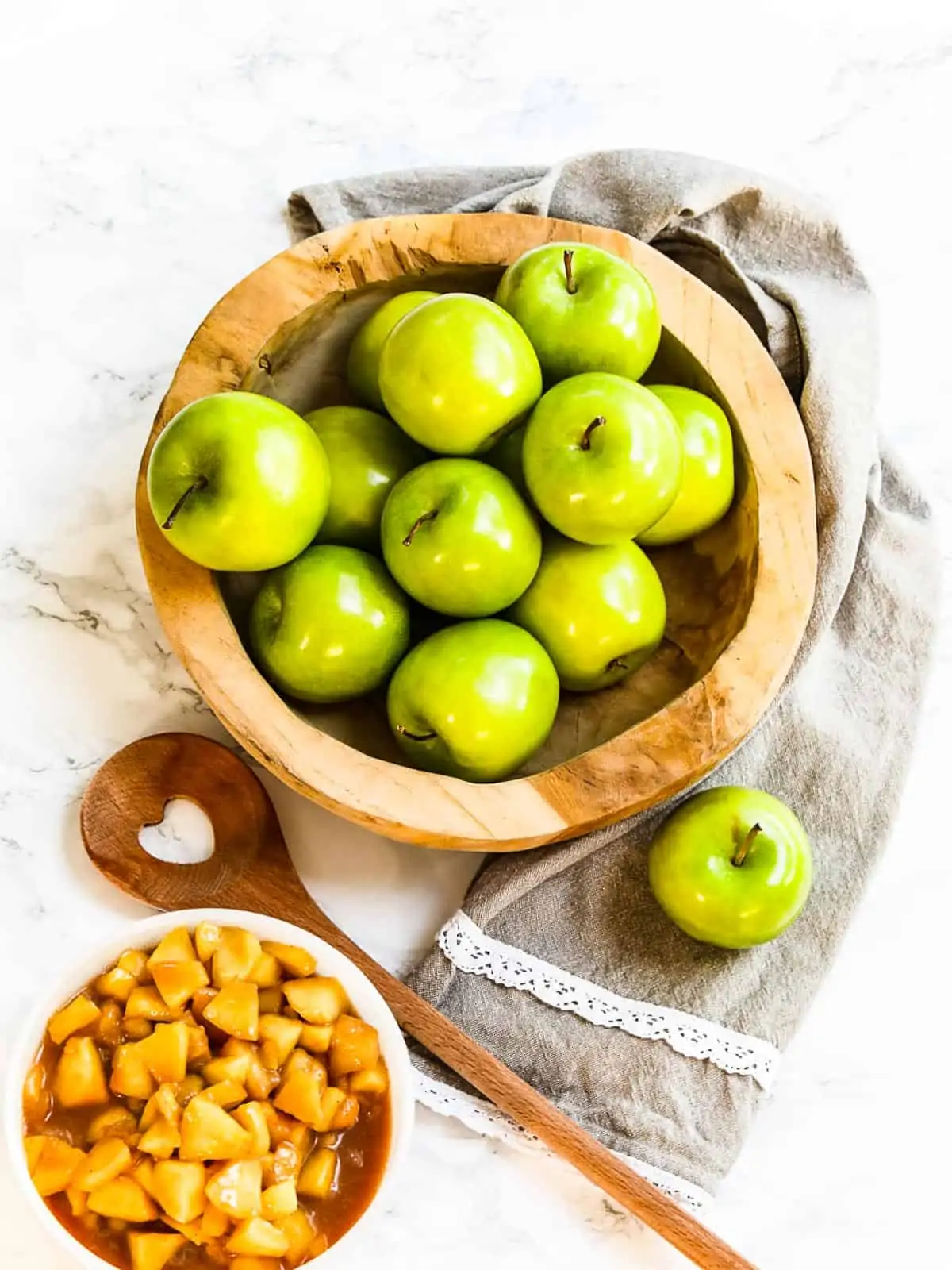 A wood bowl filled with green apples a towel, and bowl of cooked apple pie filling.