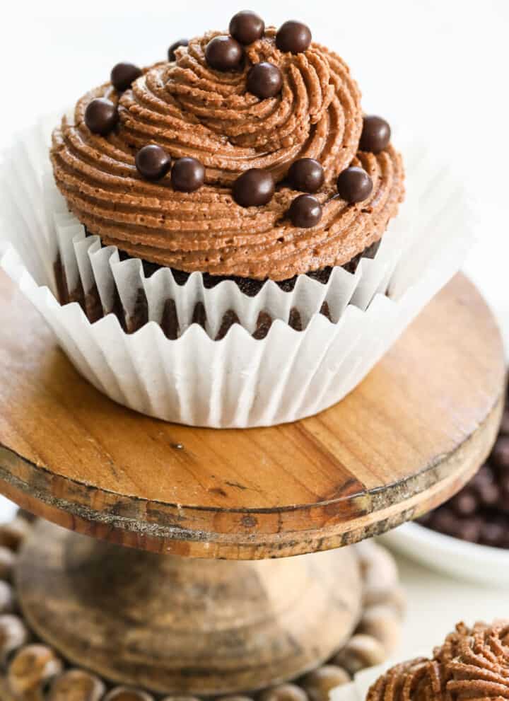 A chocolate cupcake with swirled chocolate buttercream frosting and sprinkles on a small stand.