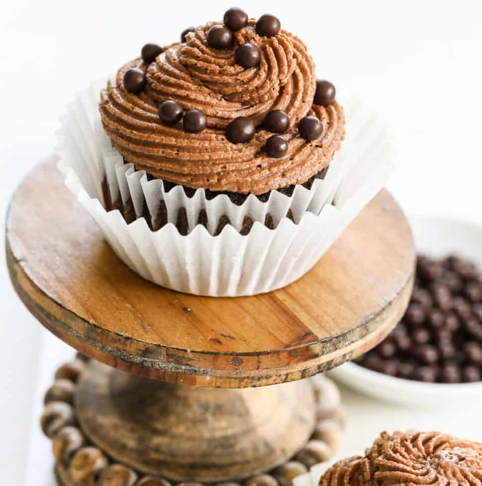 A chocolate cupcake with swirled chocolate buttercream frosting and sprinkles on a small stand.