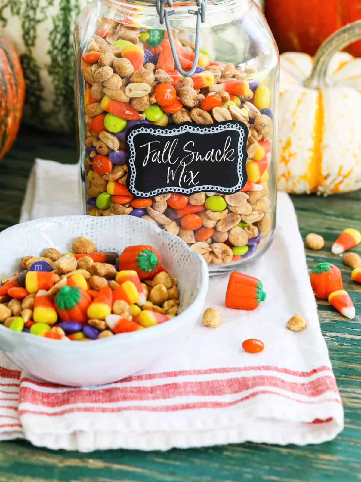 A large glass jar filled with a Fall Snack Mix with candy corn and M&Ms with a small bowl of it nearby.