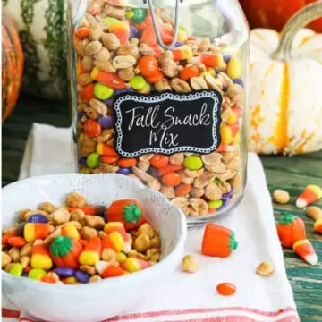 A large glass jar filled with a Fall Snack Mix with candy corn and M&Ms with a small bowl of it nearby.