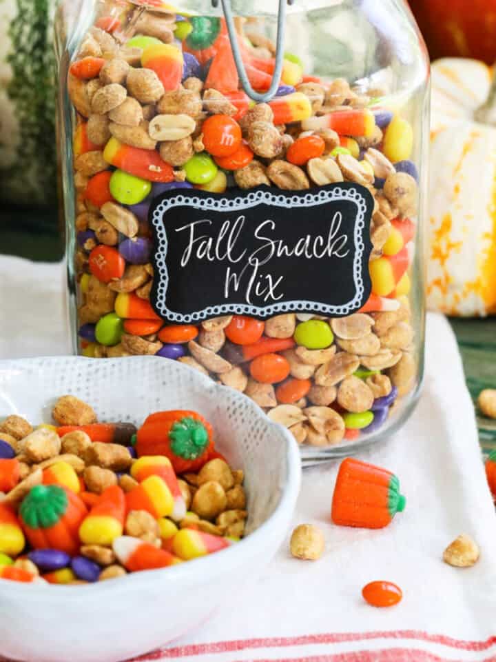 A large glass jar filled with a Fall Snack Mix with candy corn and M&Ms with a small bowl of it nearby.