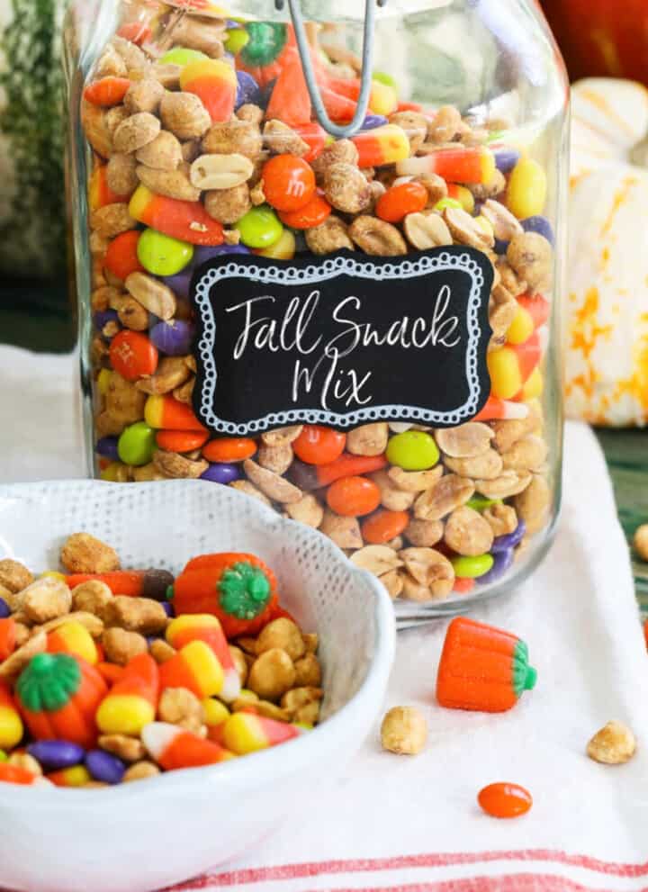 A large glass jar filled with a Fall Snack Mix with candy corn and M&Ms with a small bowl of it nearby.