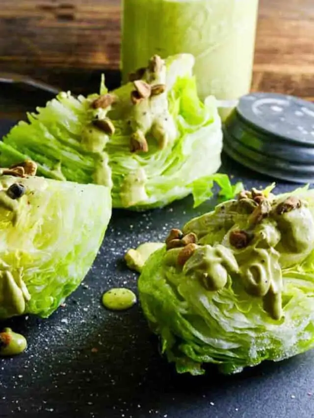 Wedge salad with avocado dressing drizzled on top with pistachios on a black slate board.