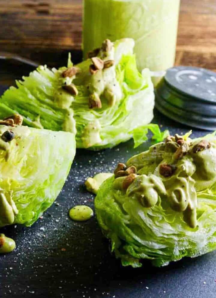 Wedge salad with avocado dressing drizzled on top with pistachios on a black slate board.