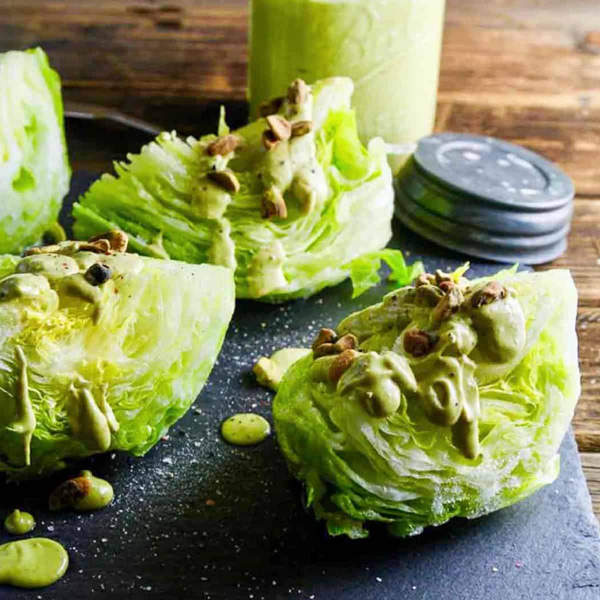 Wedge salad with avocado dressing drizzled on top with pistachios on a black slate board.