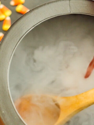 A black cauldron with wooden ladle and pouring red punch into the bowl with dry ice smoke for Halloween Punch.