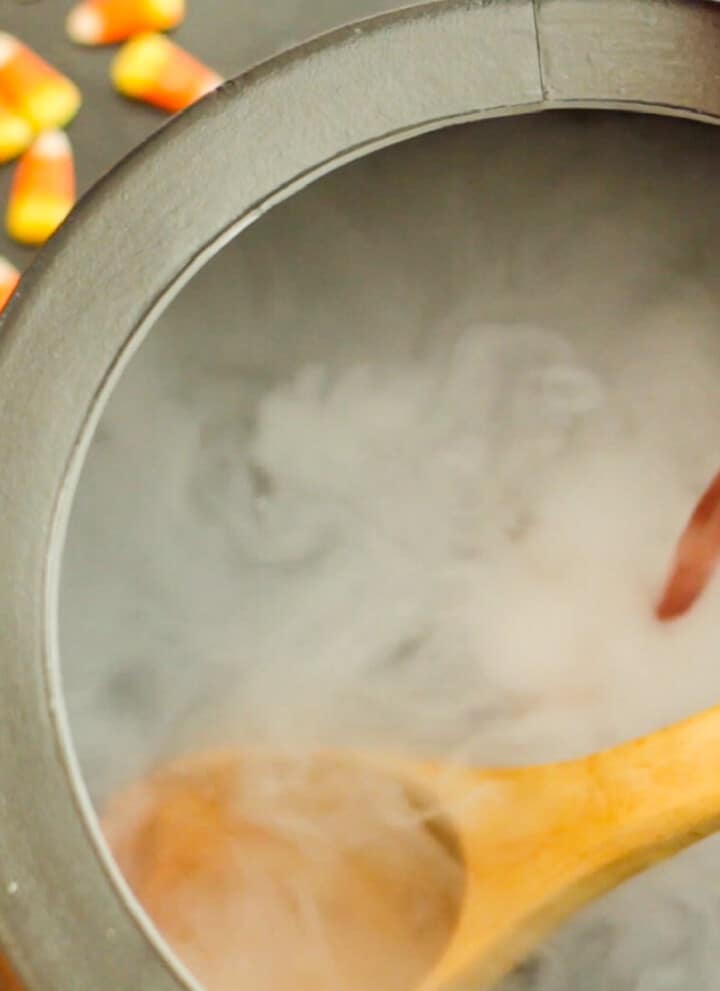 A black cauldron with wooden ladle and pouring red punch into the bowl with dry ice smoke for Halloween Punch.