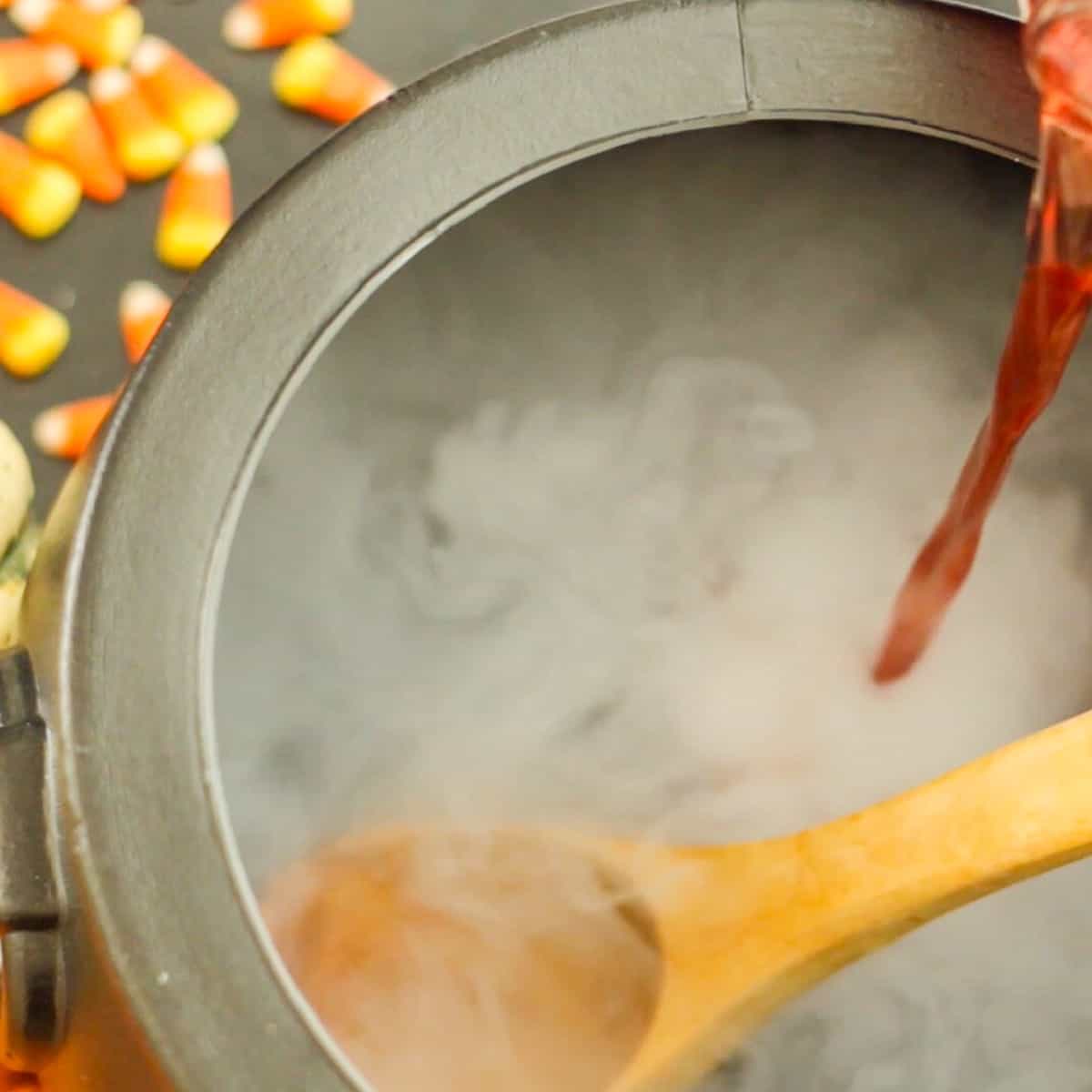 A black cauldron with wooden ladle and pouring red punch into the bowl with dry ice smoke for Halloween Punch.