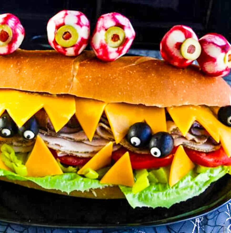 A giant sub sandwich that looks like a Monster with giant edible radish eyes and small black olive eyes.