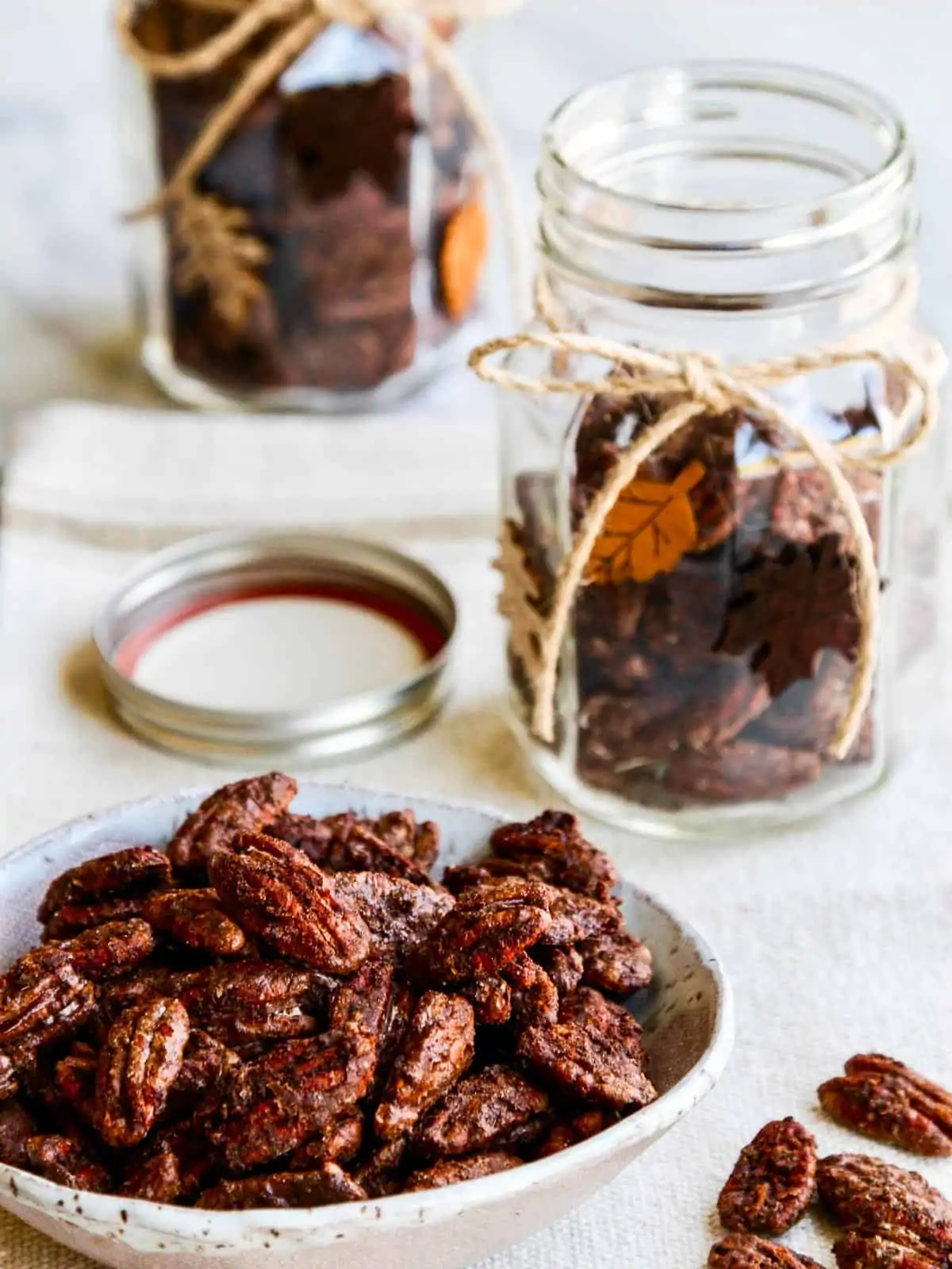 A white bowl of spicy candied pecans with two decorated gift jar nearby being filled.