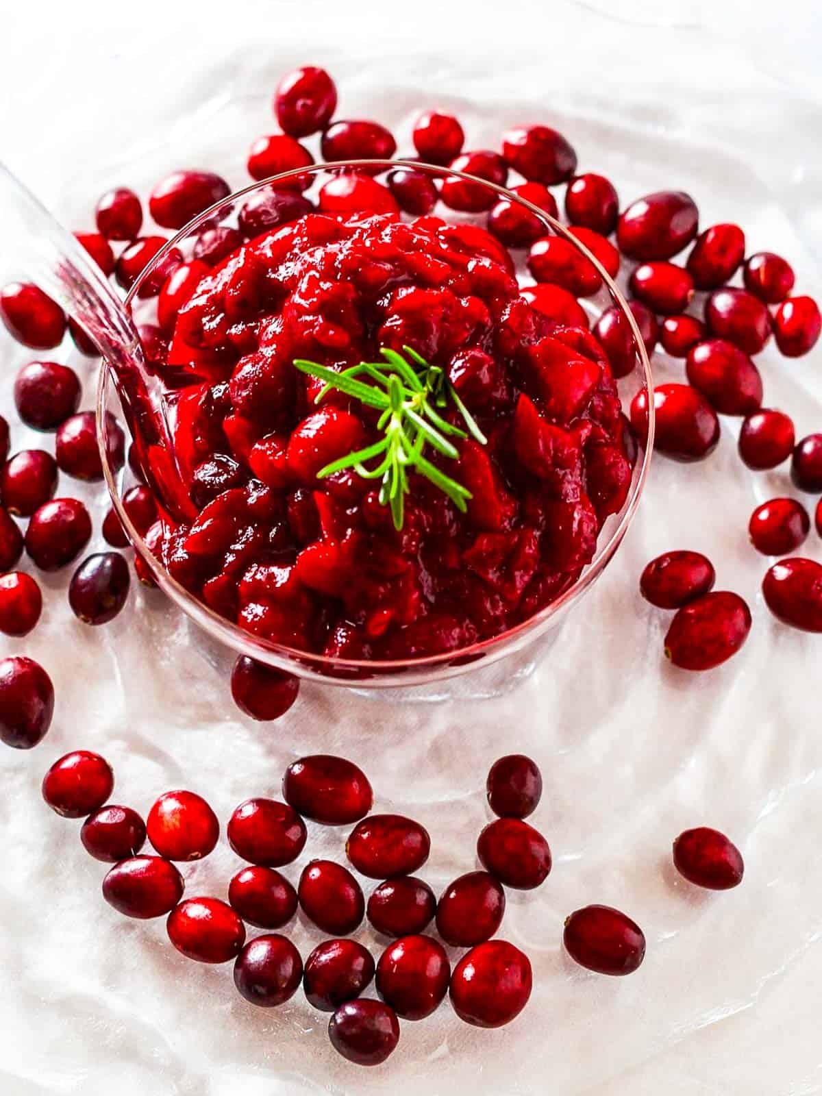 A clear glass bowl filled with pineapple cranberry orange sauce surrounded by fresh cranberries.