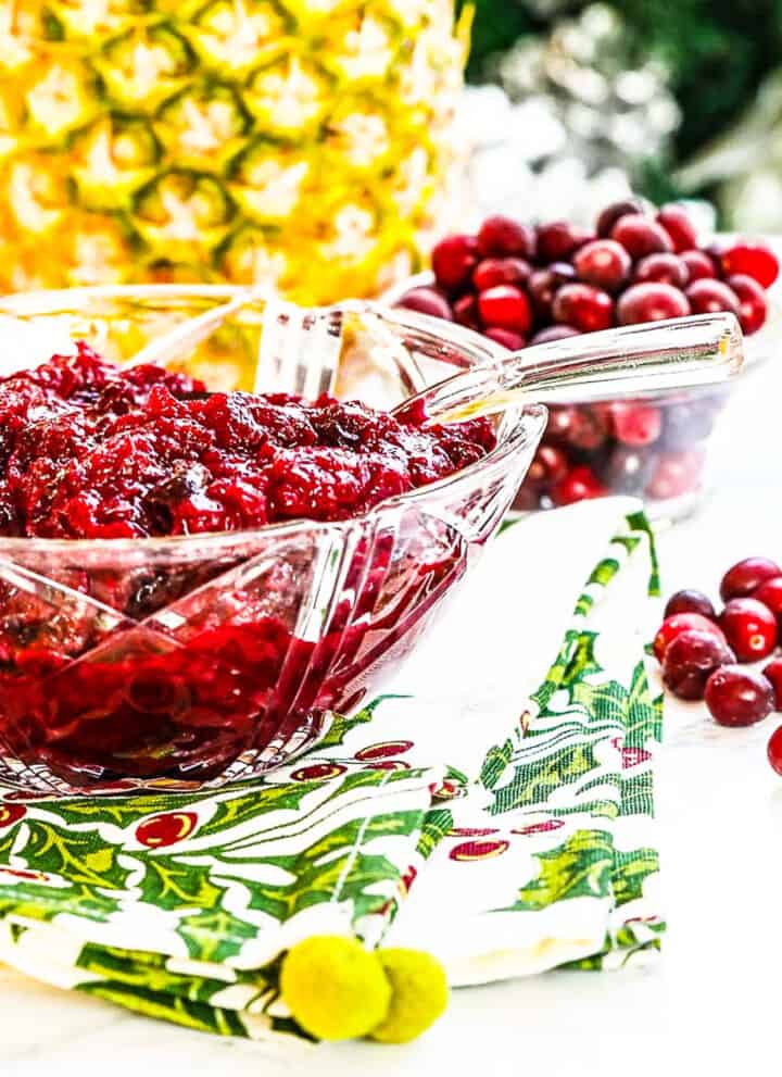 A crystal bowl filled with cranberry sauce with pineapple and cranberries in the background.