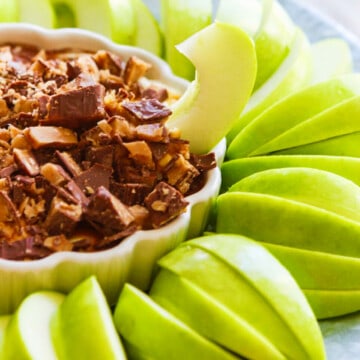 Slices of green Granny Smith apples around a white dish with a sweet dessert dip inside a low small white dish with a ruffled edge, and a piece of apple sticking into the dip.