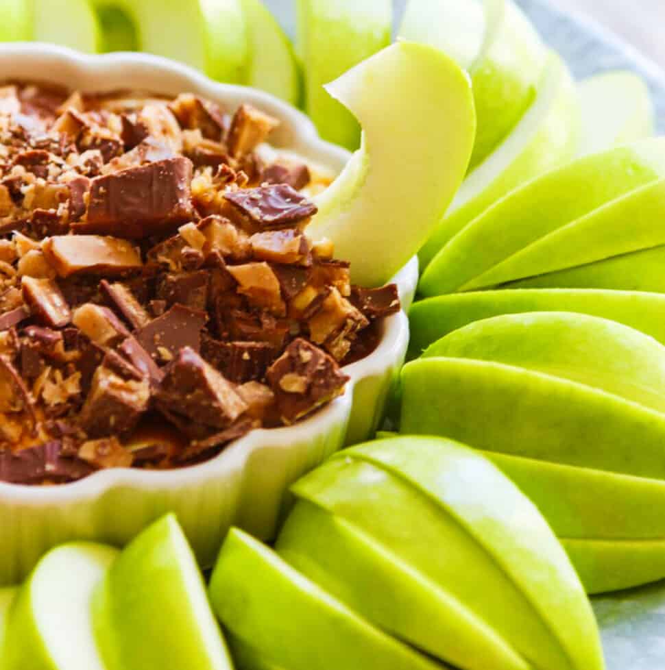 Slices of green Granny Smith apples around a white dish with a sweet dessert dip inside a low small white dish with a ruffled edge, and a piece of apple sticking into the dip.