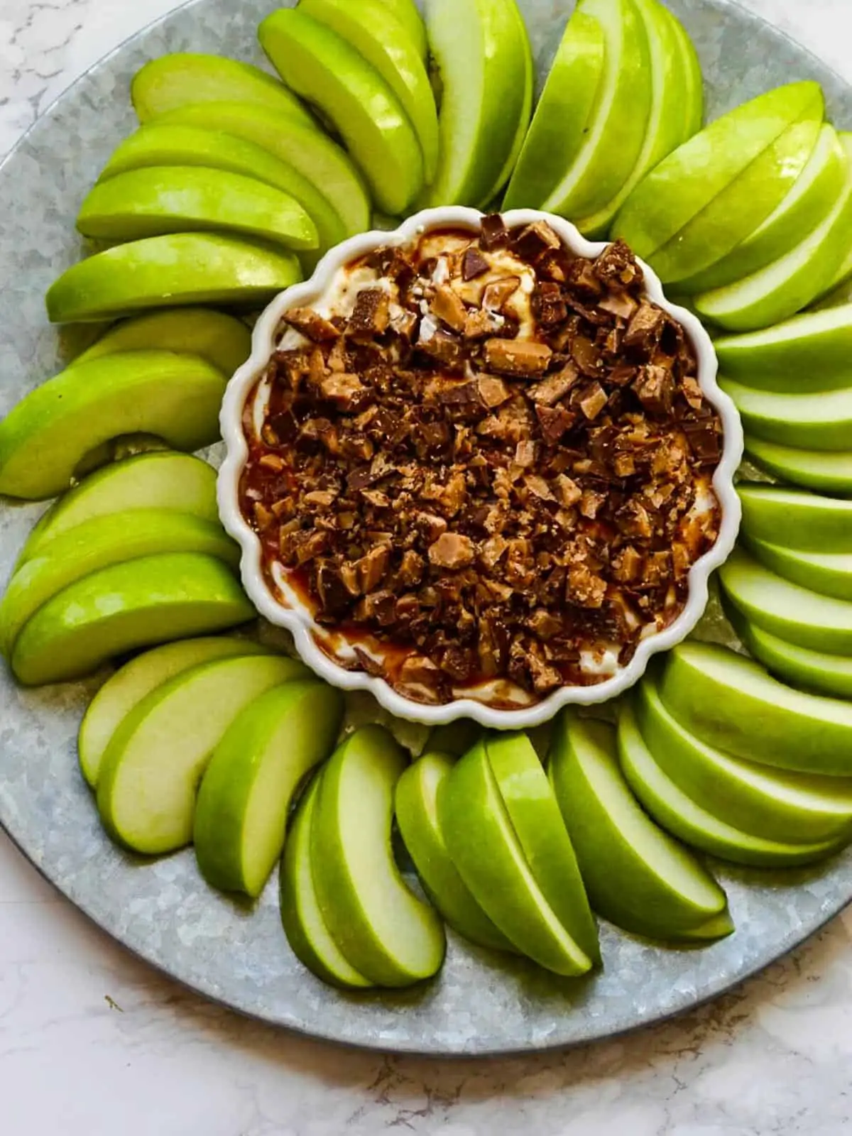 A large silver platter with green apple slices layed into a circular pattern around a white dish holding a sweet caramel apple dip for dessert topped with crushed Heath candy bars.