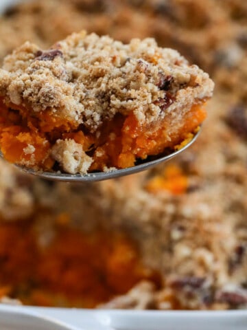 Lifting a scoop of sweet potato casserole out of a white casserole dish with large serving spoon.