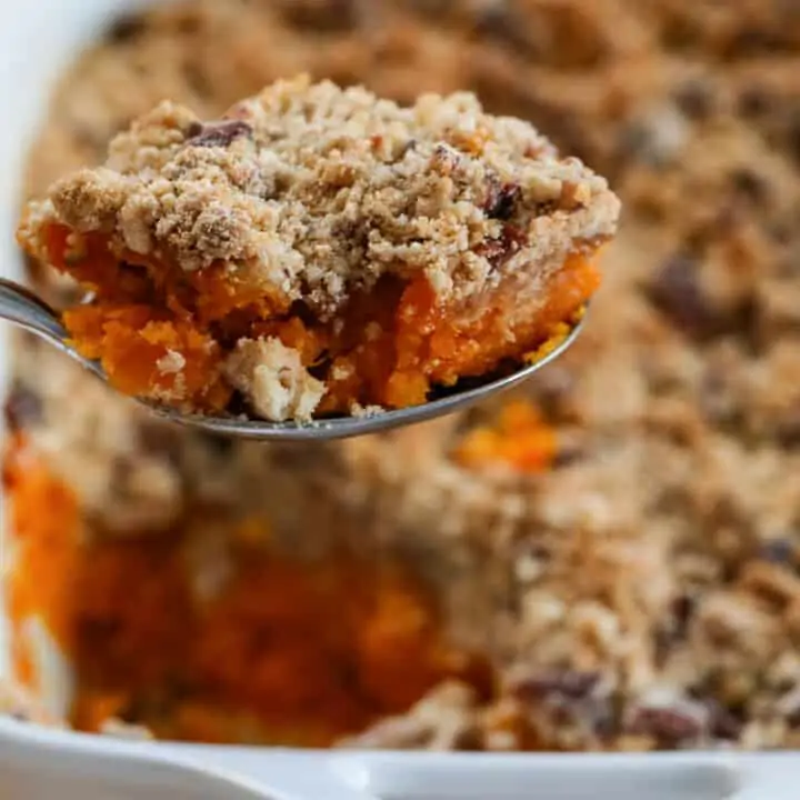 Lifting a scoop of sweet potato casserole out of a white casserole dish with large serving spoon.