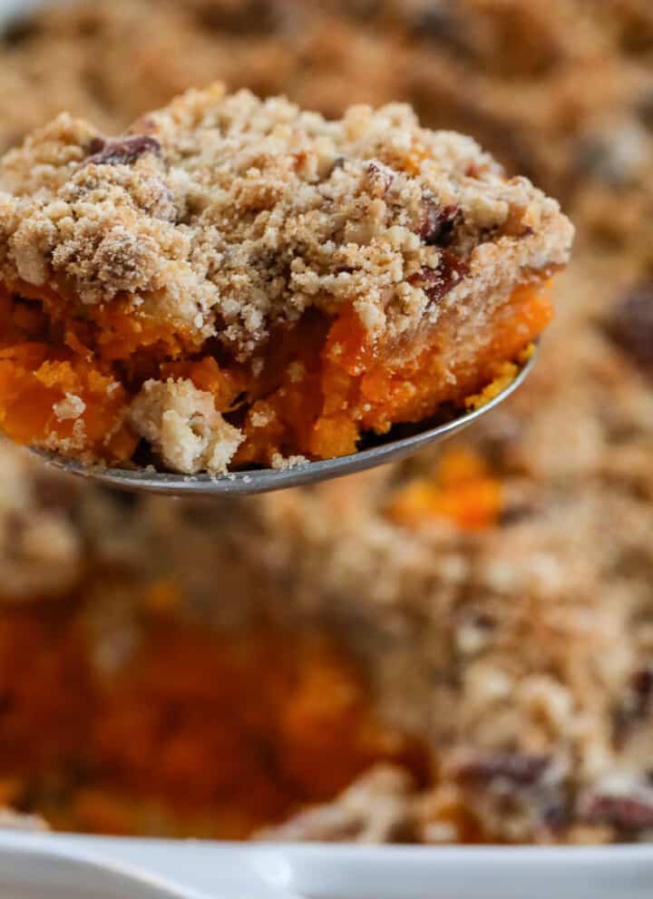 Lifting a scoop of sweet potato casserole out of a white casserole dish with large serving spoon.