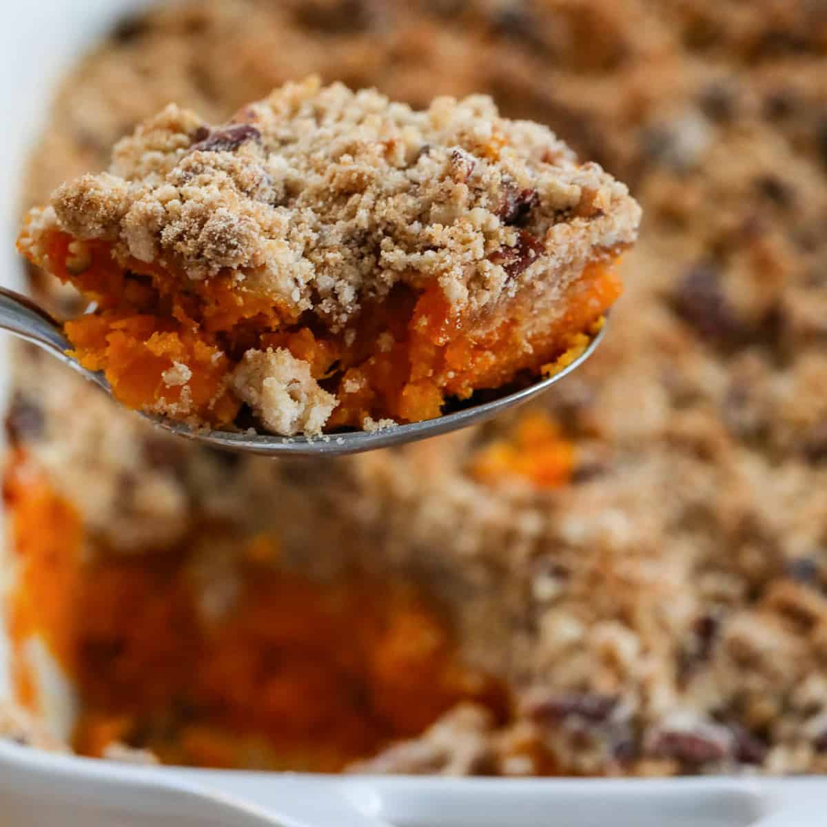 Lifting a scoop of sweet potato casserole out of a white casserole dish with large serving spoon.