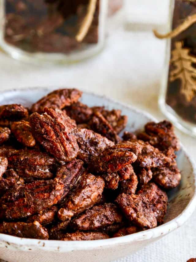 A white bowl with candied pecans and a few on the table with jars filled with them nearby.