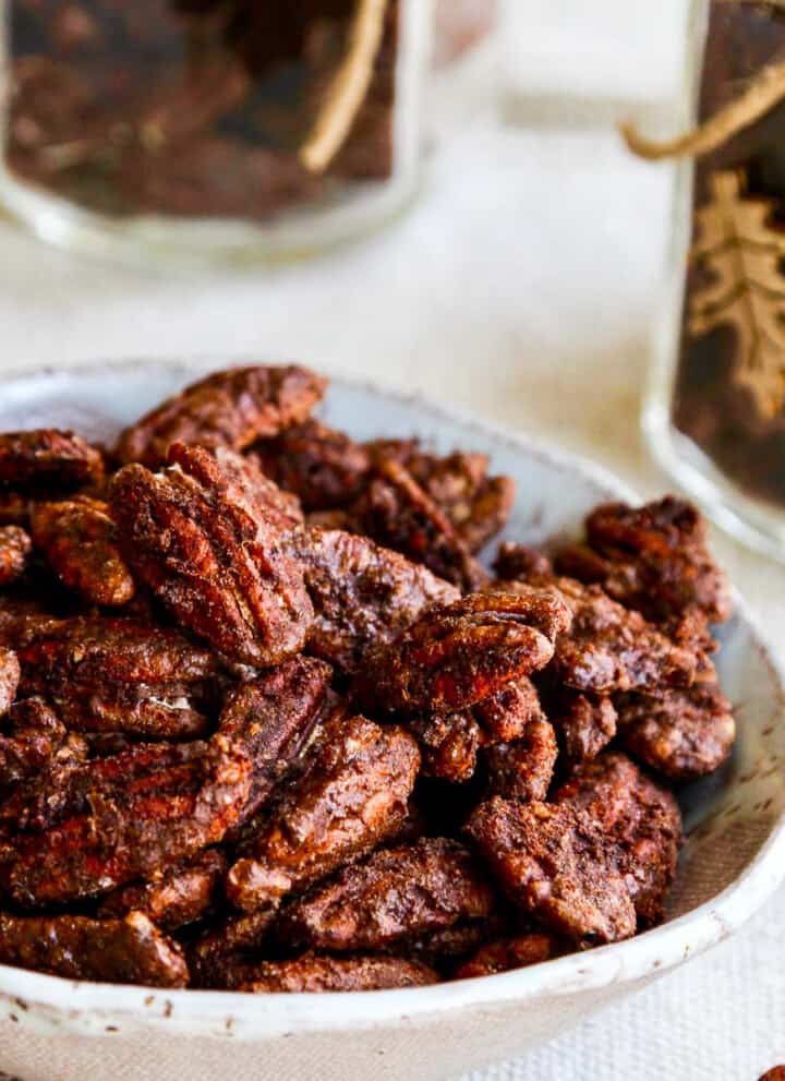 A white bowl with candied pecans and a few on the table with jars filled with them nearby.