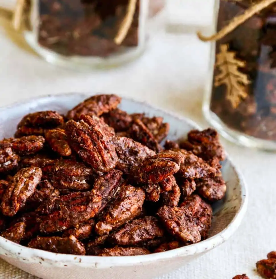 A white bowl with candied pecans and a few on the table with jars filled with them nearby.