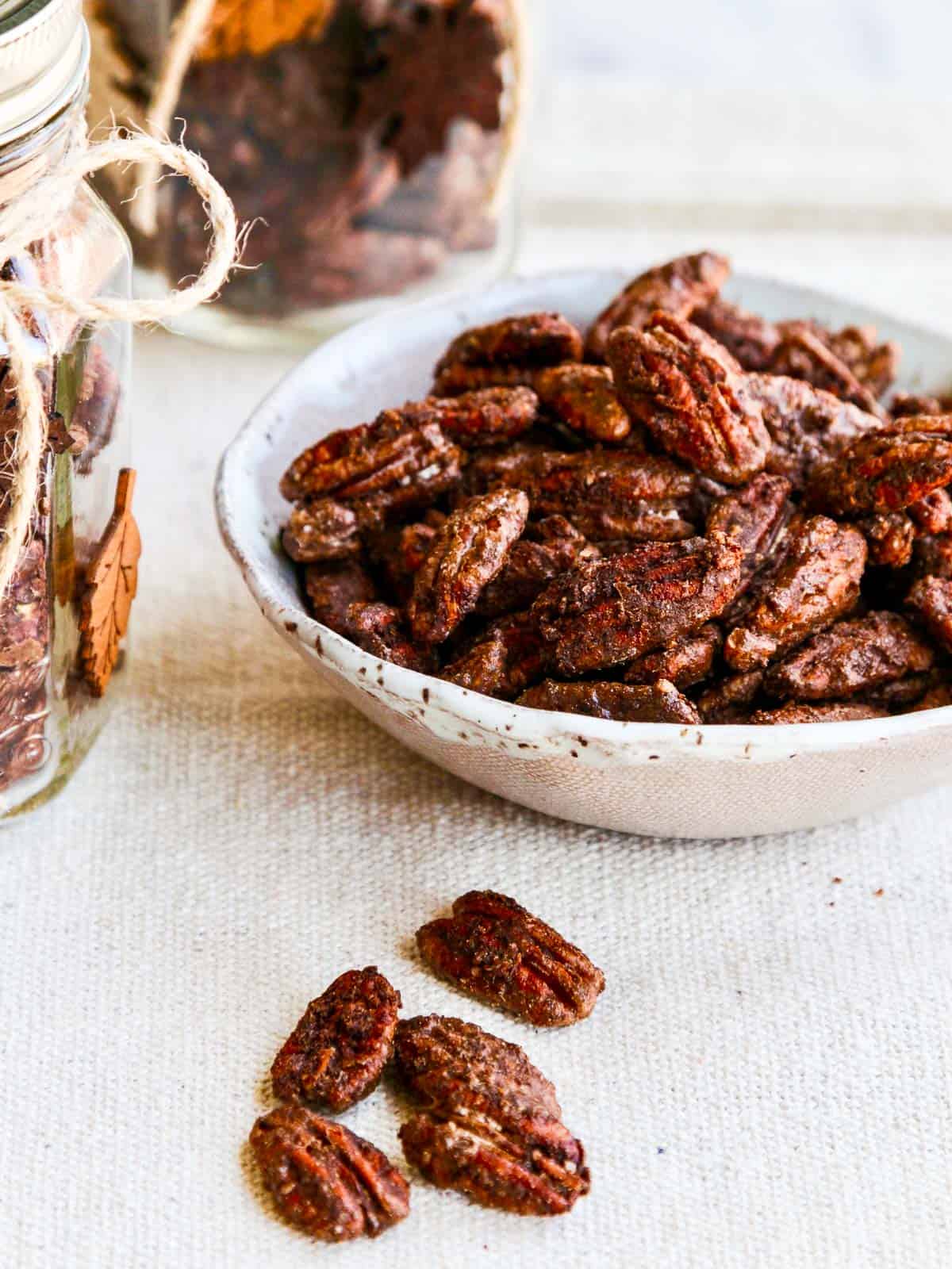 A white bowl filled with candied pecans on with a few pecans on a linen towel. 