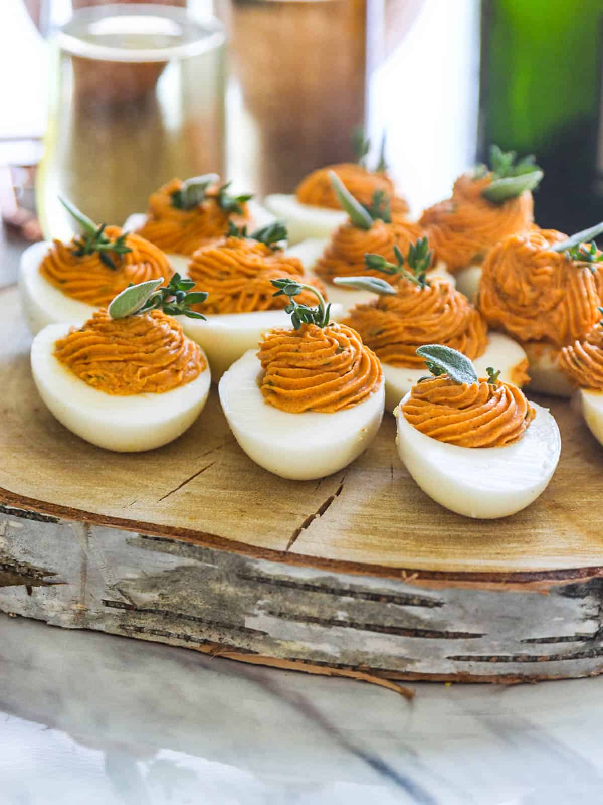 A rustic piece of tree cut into a platter with Thanksgiving deviled eggs filled with rust colored filling and garnished with herbs.