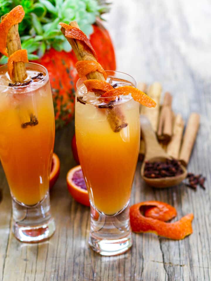 Two tall glasses of Fireball Whisky Cocktails garnished with orange peel wrapped cinnamon sticks.