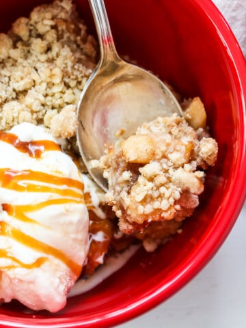 Apple Crisp in a red bowl with a silver spoon, ice cream, and drizzled with caramel sauce.