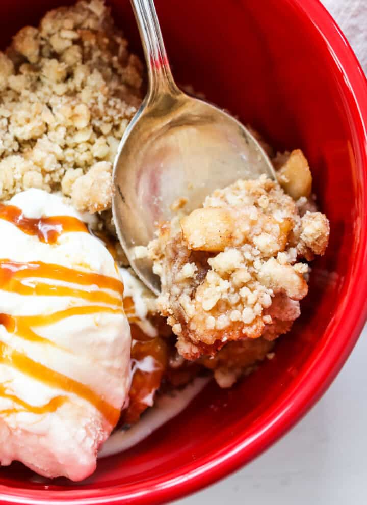 Apple Crisp in a red bowl with a silver spoon, ice cream, and drizzled with caramel sauce.