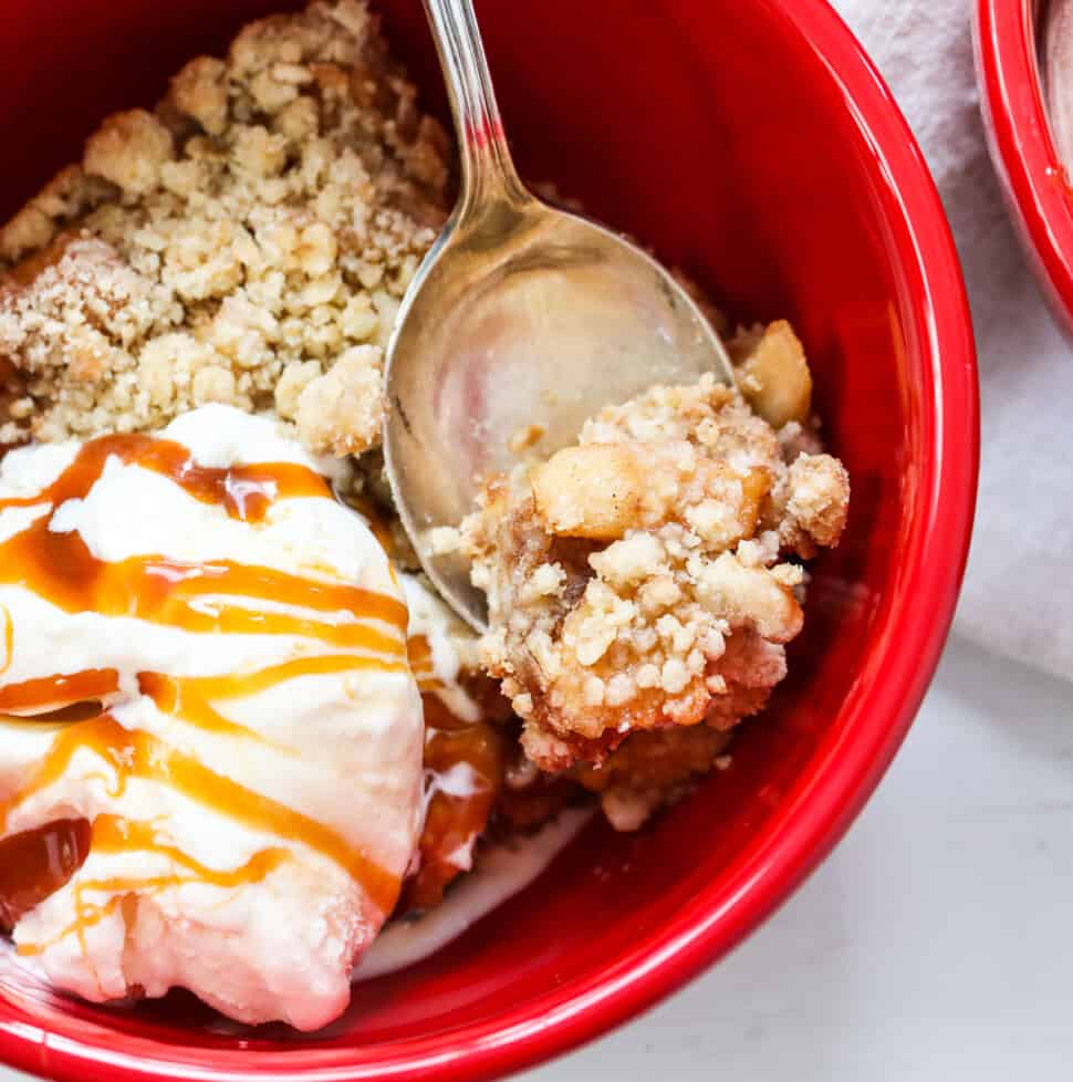 Apple Crisp in a red bowl with a silver spoon, ice cream, and drizzled with caramel sauce.