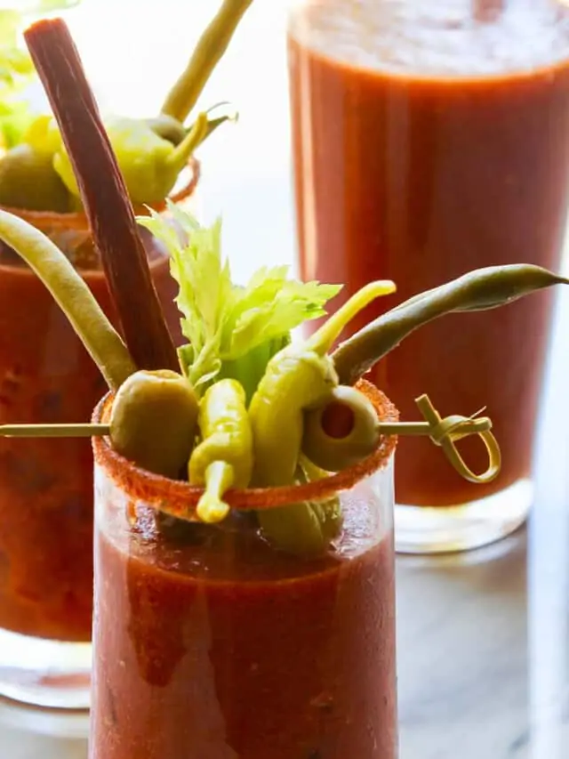 A pitcher of bloody marys with two drinks garnished with olives, pepperoncinis, and beef sticks.