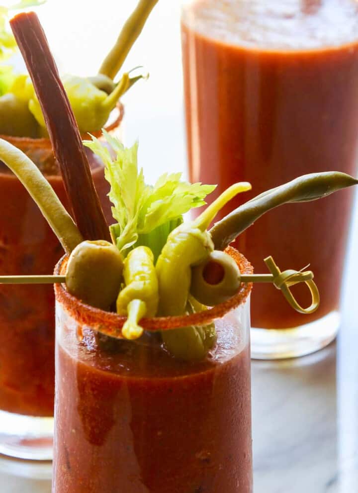 A pitcher of bloody marys with two drinks garnished with olives, pepperoncinis, and beef sticks.