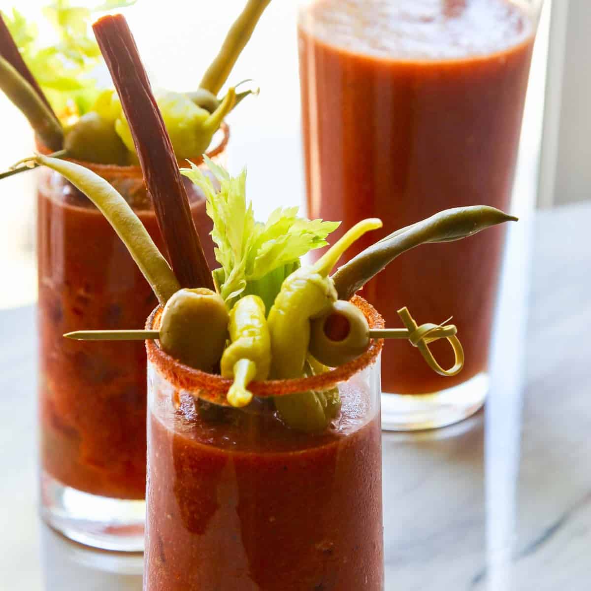 A pitcher of bloody marys with two drinks garnished with olives, pepperoncinis, and beef sticks.
