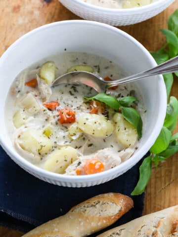 A white bowl full of Chicken Gnocchi Soup with a silver spoon and bread nearby.