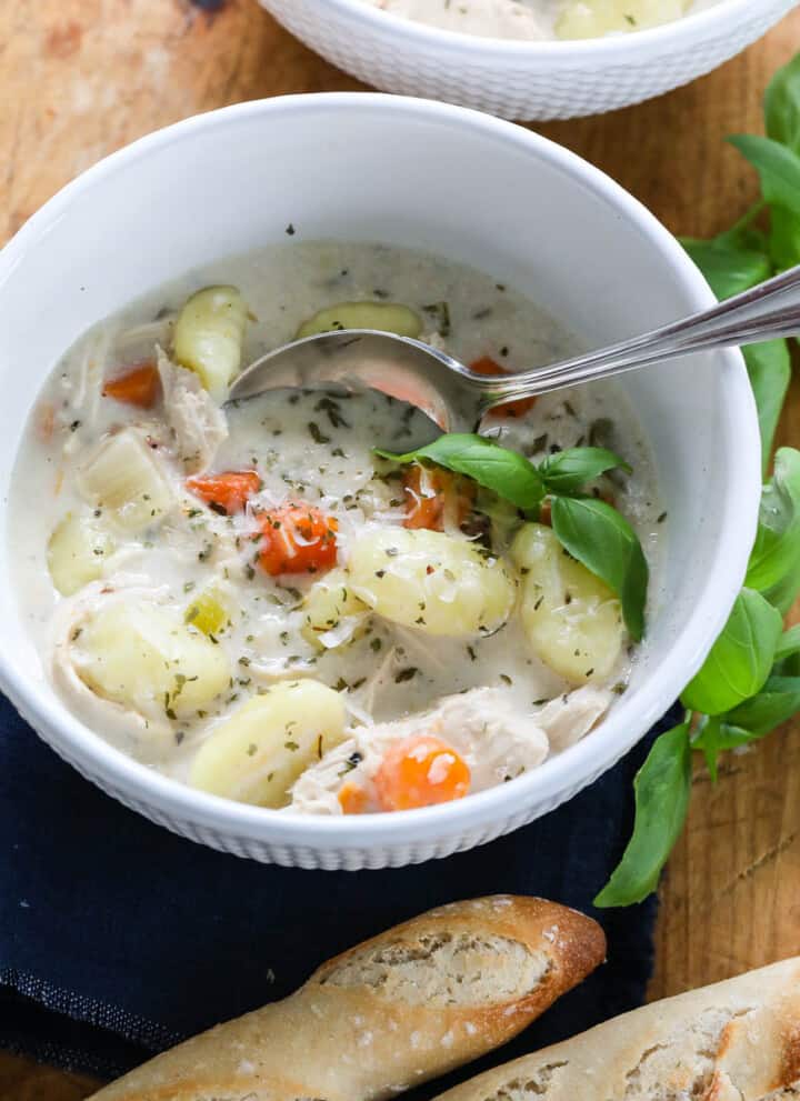 A white bowl full of Chicken Gnocchi Soup with a silver spoon and bread nearby.