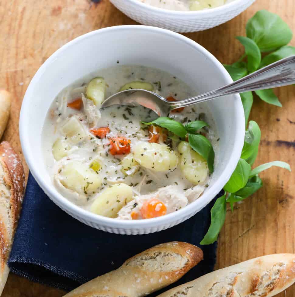 A white bowl full of Chicken Gnocchi Soup with a silver spoon and bread nearby.