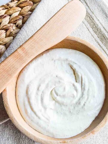 Looking down into a wooden bowl with creamy balsamic horseradish sauce swirled in the center, and a wooden spreader resting on the edge of the bowl with a rustic beige tablecloth underneath and on a woven rattan placemat.