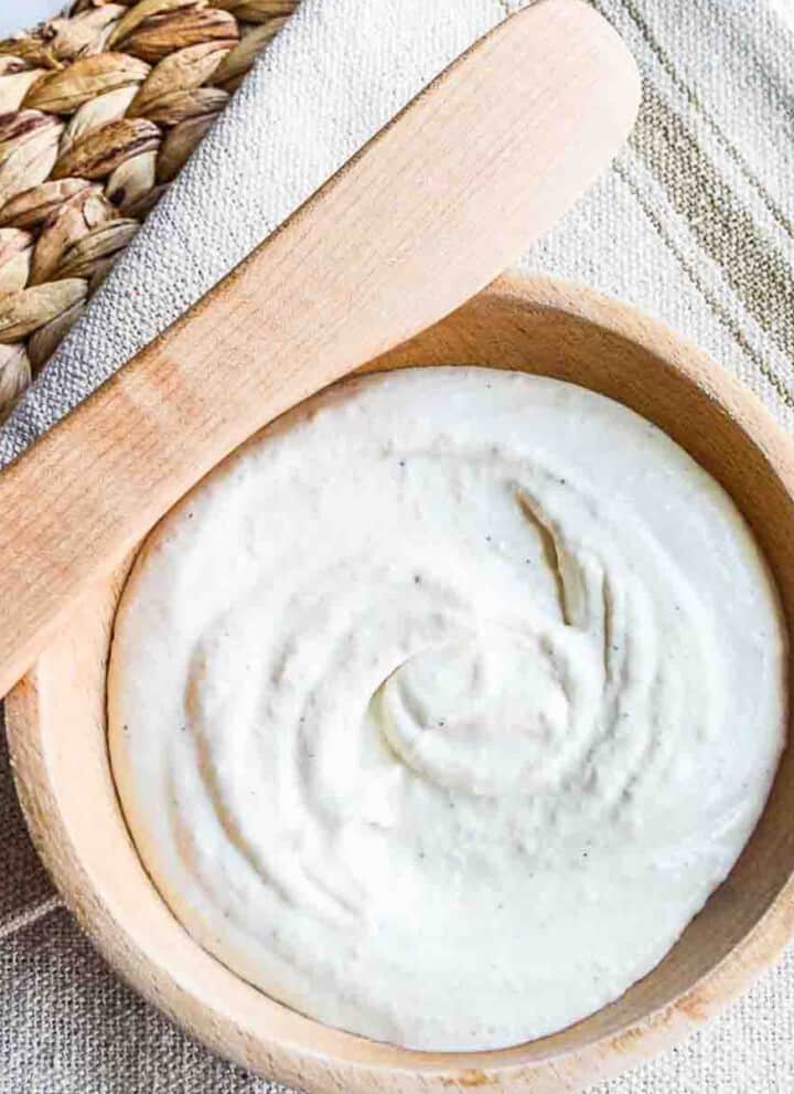 Looking down into a wooden bowl with creamy balsamic horseradish sauce swirled in the center, and a wooden spreader resting on the edge of the bowl with a rustic beige tablecloth underneath and on a woven rattan placemat.