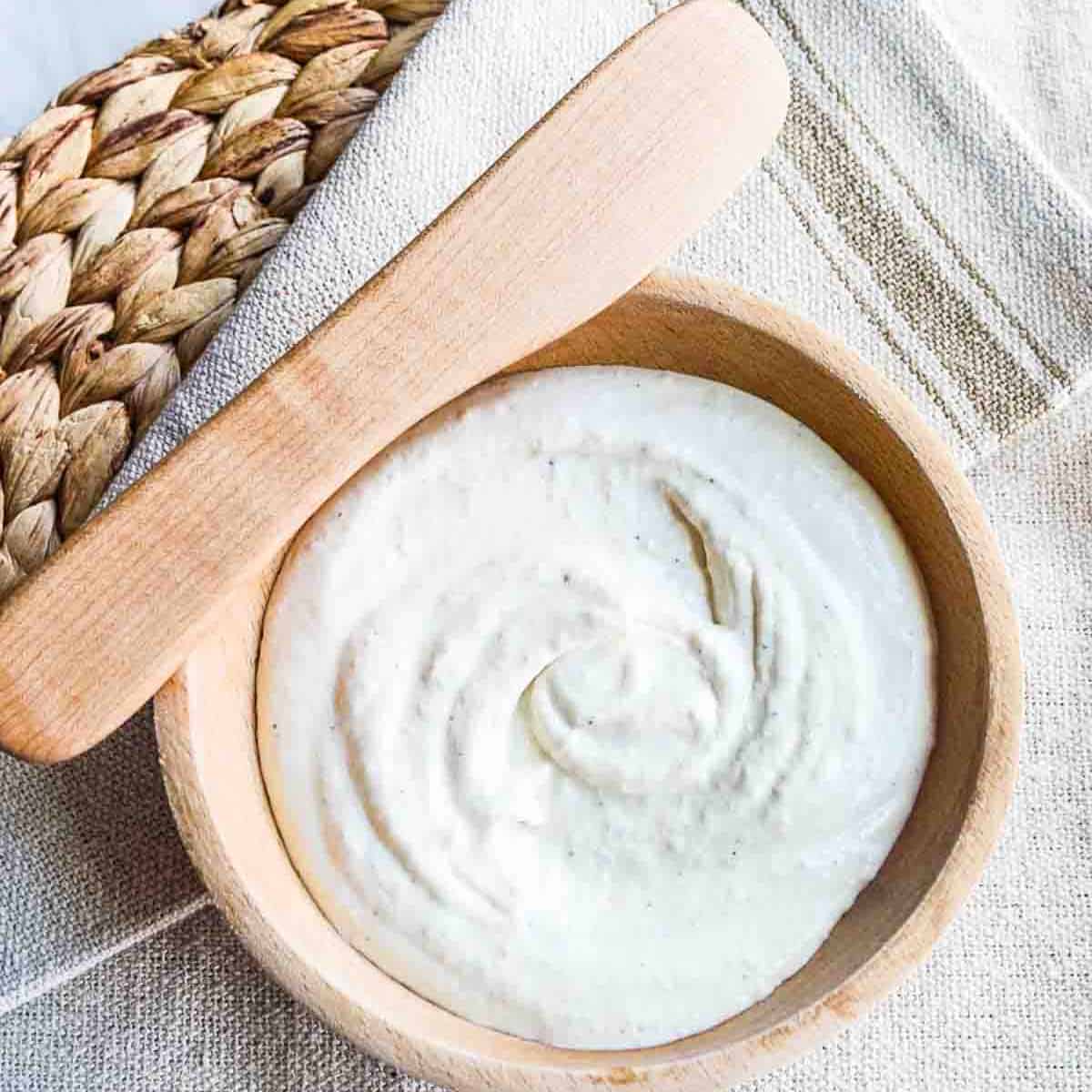 Looking down into a wooden bowl with creamy balsamic horseradish sauce swirled in the center, and a wooden spreader resting on the edge of the bowl with a rustic beige tablecloth underneath and on a woven rattan placemat.