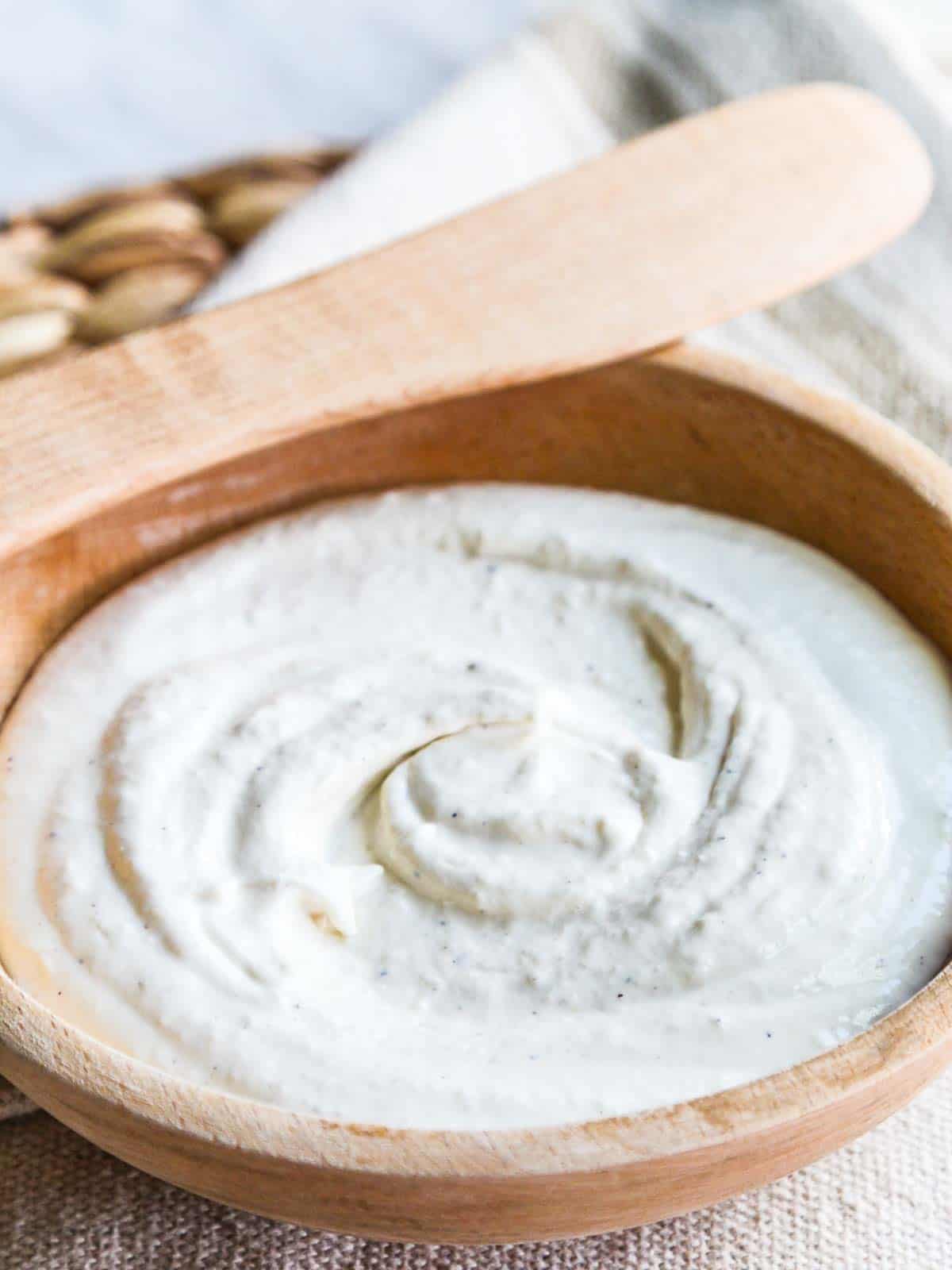 A wooden bowl with creamy balsamic horseradish sauce swirled in the center, and a wooden spreader resting on the edge of the bowl with a rustic beige tablecloth underneath.