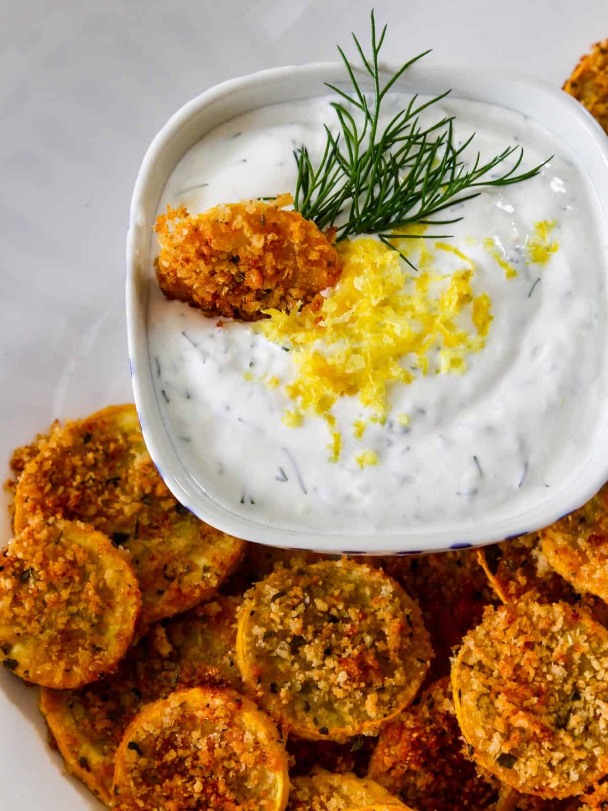 A zucchini chip stuck in a small white bowl of Lemon Dill yogurt dip garnished with dill and lemon.