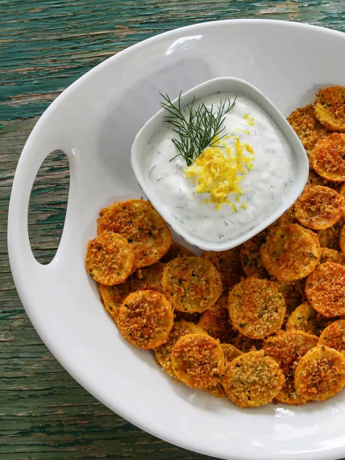 A large white platter with zucchini chips on it and a small bowl of yogurt dip garnished with lemon and dill.